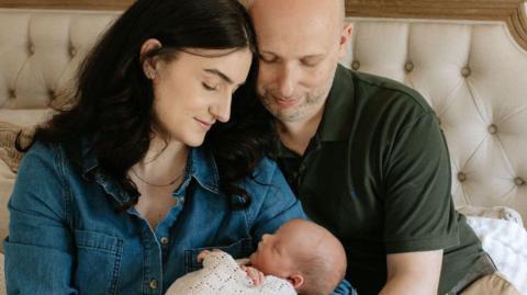 A woman with black hair and denim shirt holds a baby wrapped in a white blanket while a man in a green polo looks down