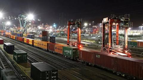 Southampton Docks at night with a freight train loaded with containers in the foreground extends back into the distance. Floodlights illuminate stacks of freight containers are piled up in the background.