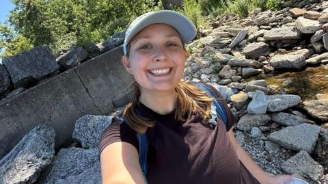 Megan Szostak stands in a rocky stream, taking a selfie and smiling at the camera. She wears a black t-shirt, blue backpack and blue hat.