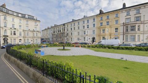 The Crescent in Bridlington. It shows a green area surrounded by black railings with pavement in the middle of the grass. A road surrounds the green area and there are residential flats.