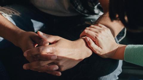 Generic close up image of a man sitting down with his fingers interlocked while a woman rests her hand on his forearm in a supportive way