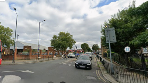 Google street view image of Pound Lane from its junction with High Road, showing a road with several cars, pedestrians and what looks like a large grocery store to the left.