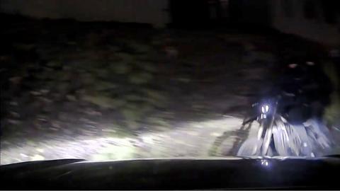 Two masked men on a black motorbike, both wearing all black, illuminated by car lights, riding next to a hedge.