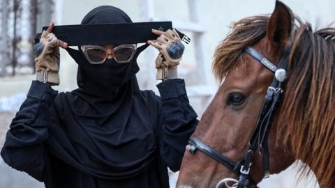 Woman wearing sunglasses and Islamic dress - including an abaya and niqab - gets ready to mount her chestnut-coloured horse.