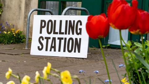 A polling station sign seen through a row of colourful Spring flowers in the foreground.