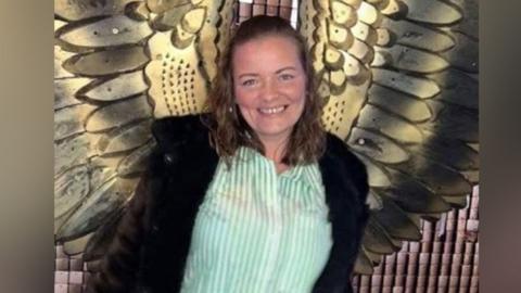 Dianne Cleary pictured in front of a wall with a large bronze set of angel wings on it. She is smiling and wearing a stripey green and white shirt and open black coat. Her wavy hair is half pinned back from her face.