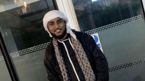 Ahmed Ali Jama is seen wearing a white headscarf and a patterned scarf over a black jacket stands smiling in front of a glass building entrance.
