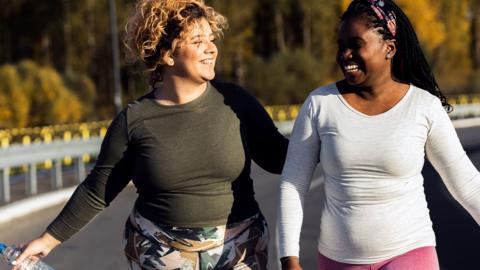 Two women in sports clothes walking on a road, one holding a water bottle