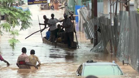 Men are using a canoe to navigate a road that has turned into a river. Other people are wading through brown floodwater and a car is nearly submerged.