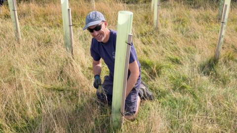 NECF Forest Manager Lloyd Jones crouches behind a sapling that has just been planted. He is wearing a dark t-shirt and shorts, baseball cap and sunglasses and is smiling. The sapling has a semi-translucent, green plastic tree guard around it and in the background there are several other newly-planted trees.