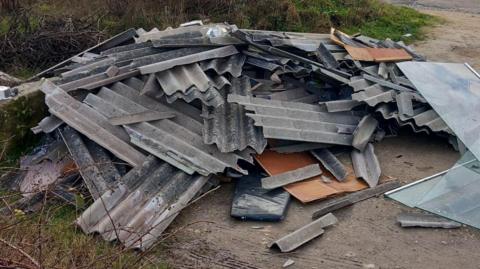 Image of a large amount of fly-tipped, corrugated asbestos sheets and large panes of glass. In the background can be seen a rough track and grass embankments