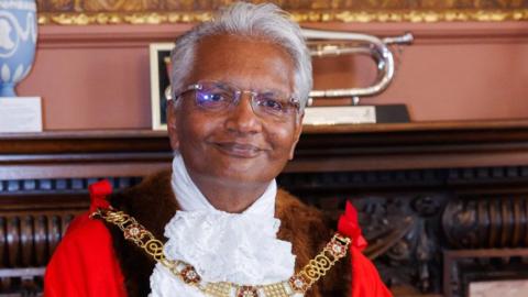 Wearing his mayoral robes and gold chain, Bharat Pankhania smiles at the camera. Behind him two vases and a musical instrument can be seen on a mantelpiece.