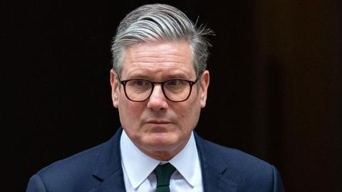 Sir Keir Starmer looks to the left of the camera while wearing suit, tie and glasses as he comes out of 10 Downing Street in London