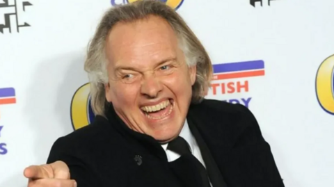 Rik Mayall, who has shoulder length hair has a huge grin on his face, standing against a white backdrop at an awards ceremony. He is wearing a black suit and tie with a white shirt and is pointing with his right index finger.