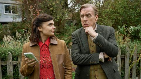 Gwyneth Keyworth and Timothy Spall as their Death Valley characters John and Janey. They look at each other and stand in front of a garden fence. 