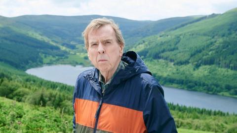 Timothy Spall in a waterproof jacket standing in the Bannau Brycheiniog/Brecon Beacons. There is a lake or reservoir behind him and hills around.