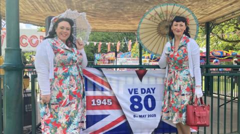 Two women in floral dresses and white cardigans stand next to a bandstand in a park. They are wearing white cardigans and holding sun umbrellas.