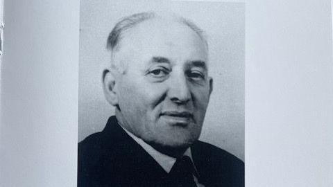 A black and white photo of an older man, with short, balding hair. He is wearing a white shirt and black suit. 