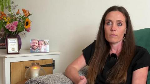 Victoria Rodwell is sitting on a sofa at home. She has long brown hair and is wearing a black T-shirt. A candle is burning next to her and there is a vase of flowers on a cupboard, with a certificate from her 2022 Channel swim and her engagement picture.