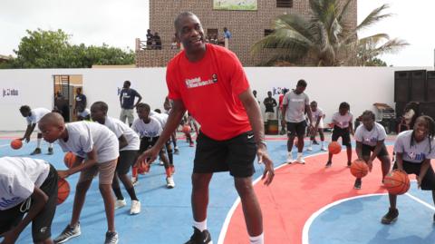 Dikembe Mutombo in shorts and t-shirt smiles as he leads a basketball class for young children on a court in Africa