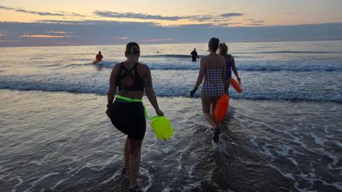 Swimmers wearing swimming costumes walk out into the sea at Scarborough, holding bright buoyancy aids.
