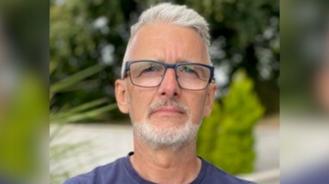 Charles Cowan, he has short grey hair and a beard. He is wearing a blue T-shirt and glasses.