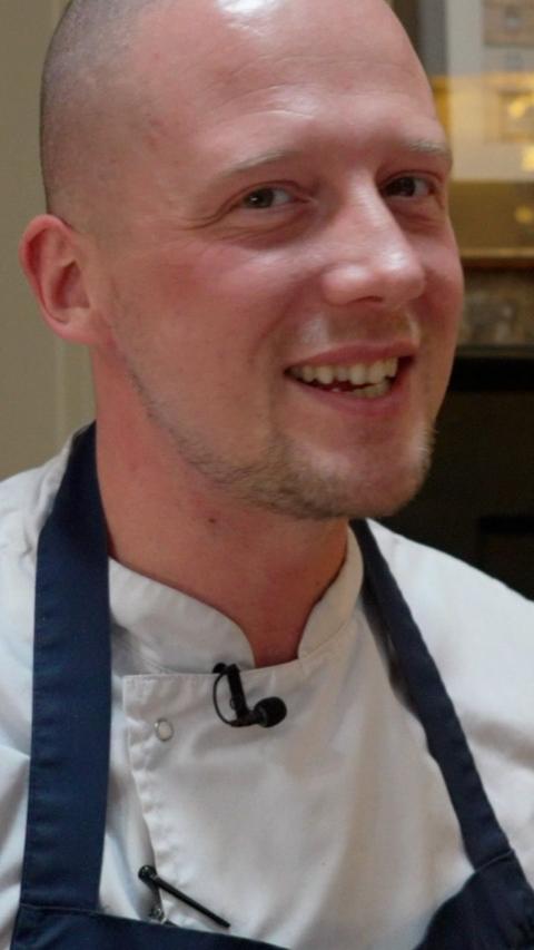 Luke Emmess grins at the camera. He is wearing chef whites and a blue apron over the top. He has light stubble on his chin and a shaved head.