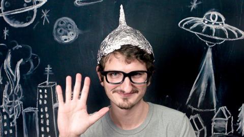 A man wears a tinfoil hat and waves at the camera, surrounded by chalk images of a UFO flying over buildings