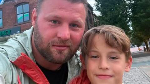 Shaun has short brown hair and a short brown beard. He is wearing a grey coat with red bits. His son has floppy brown hair and is smiling. They are standing in front of a brick building and several trees.