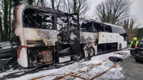 A white National Express coach has been severely damaged by a fire. The vehicle is badly burnt and charred with a number of its windows and pieces of panelling destroyed. A firefighter in a high-vis jacket and helmet is standing next to the coach.