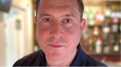 A close-up shot of Michael Westwood. He is wearing a navy blue T-shirt and looking straight into the camera lens with a neutral expression on his face.