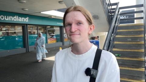 A young white man with reddish blonde hair tied back smiles at the camera. He is in the street, and wears a white top. A branch of Poundland is pictured behind him, as is a concrete flight of stairs.