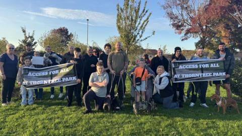 About two dozen people are standing in a park and facing the camera, with some pulling defiant poses. They are holding up signs that say: "Access for all at Thorntree Park".