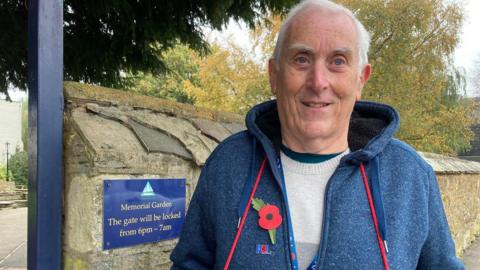 An older man standing outside a memorial garden with a poppy on his blue fleece jacket and the red string holding his tray of poppies around his neck