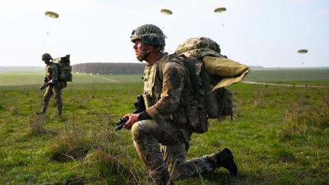 British soldier in in camouflage clothing kneeling on grass during a training exercise in England. He is holding a gun and wearing a large backpack and parachutes can be seen in the background
