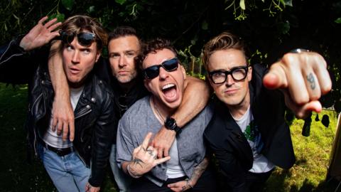 McFly, four band members, in a row. Dougie, is to the left, with sunglasses raised, with his hand on his glasses, he is wearing blue jeans, a white T-shirt and black jacket, Harry,  has his around round two men, Fanny, has two fingers raised, is wearing blue sunglasses, a blue top and his mouth open, Tom is pointing his finger, and is wearing a T-shirt and jacket. 