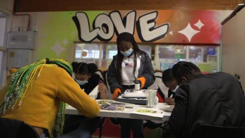 Four students are sat at a desk drawing while a woman stands in the middle of the picture working a machine that prints pictures onto t-shirts. In the background is a big Love sign written in graffiti-style lettering