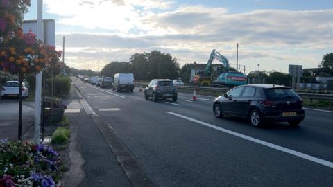 Cars are lined up on a double-carriageway road which has been reduced to one lane, with orange and white cones blocking off the other lane