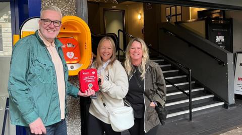 Three people with a defibrillator cabinet