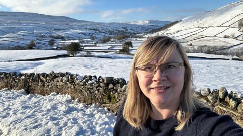 Claire Calvert on a snowy moor with dry stone walls and dales behind her covered in snow. She is a blonde woman wearing a navy jumper and glasses.