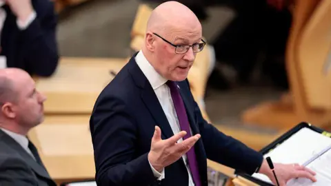 Swinney, a bald man with glasses, is wearing a dark suit jacket, white shirt and purple tie as he answers a question in the Scottish Parliament