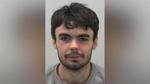 Mugshot of Evan Lyons. He has scruffy black hair and a black beard, with his right eye wider open than his left.