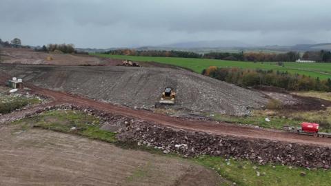 An aerial view of Flusco landfill site in Cumbria. A large mound is in the centre of the picture and two diggers are on its slopes. Beyond the site are green fields and trees.