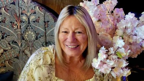 A picture of a lady with blonde hair, smiling and looking at the camera. She is wearing a yellow dress, while sat on a chair next to flowers.