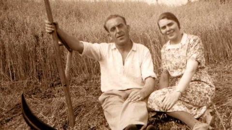 Fear le speal na làimh 's e na shuidhe air dlòthan coirce air am buain 's boireannach na suidhe ri thaobh. Coirce fhathast ri bhuain air an cùlaibh.