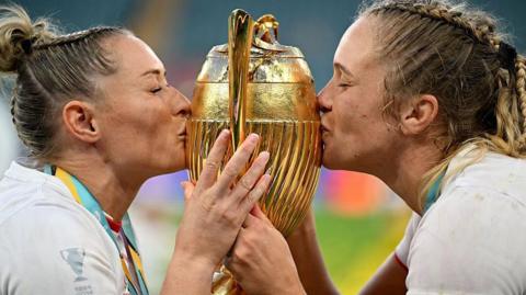 Two female rugby players, wearing white England kits and with medals around their necks, are either side of a gold trophy kissing it.