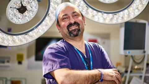 Dr Zaed Hamady smiles at the camera in an operating theatre. He has a goatee beard and wears a purple medical gown as well as a head cover
