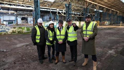 Four men and one woman wearing hi-viz jackets, boots and hard hats stand on a building site and smile at the camera