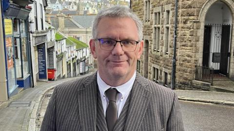 Johnnie Wells wearing a brown pin striped three-piece suit with a brown tie. He has short grey hair and is wearing a pair of glasses. He is standing at the top of Tregenna Hill. 