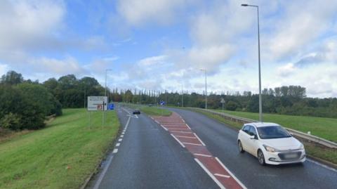 Cars are seen travelling along a road with a traffic island in the middle for a junction and the accompanying signs. The road is lined by grass banks and trees.